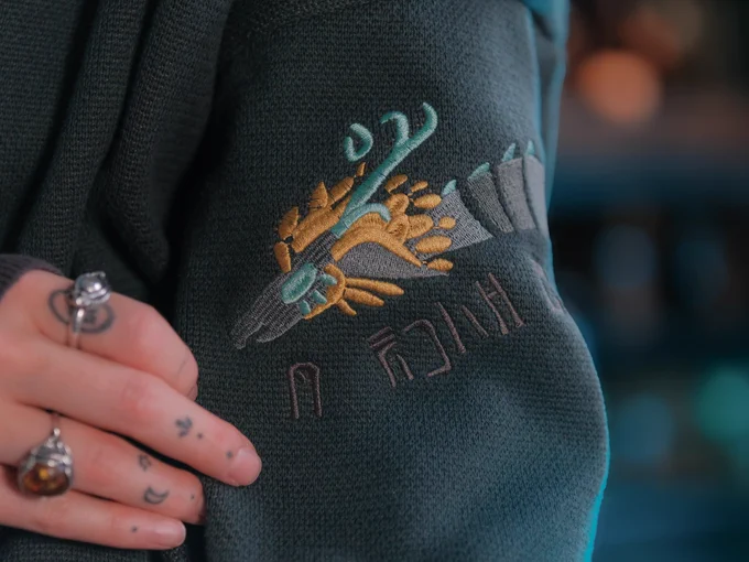 close up of persons arm wearing a cardigan with video game inspired design, a dragon, archaic style, embroidered and jacquard knit, made by knock thrice. Person is holding a sword and wearing  rings