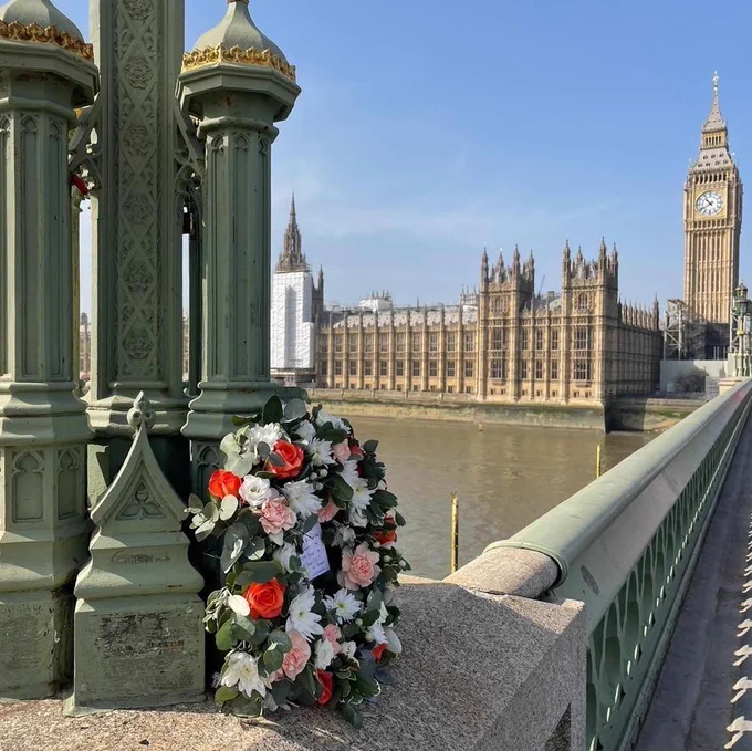 An image showing a commemorative wreath laid at Westminster Bridge, with the Thames River and Palace of Westminster visible in the background.