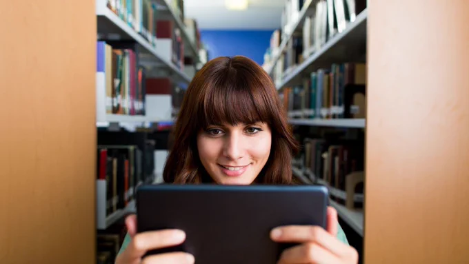 Smiling student reading on a tablet in a library, surrounded by bookshelves filled with books.