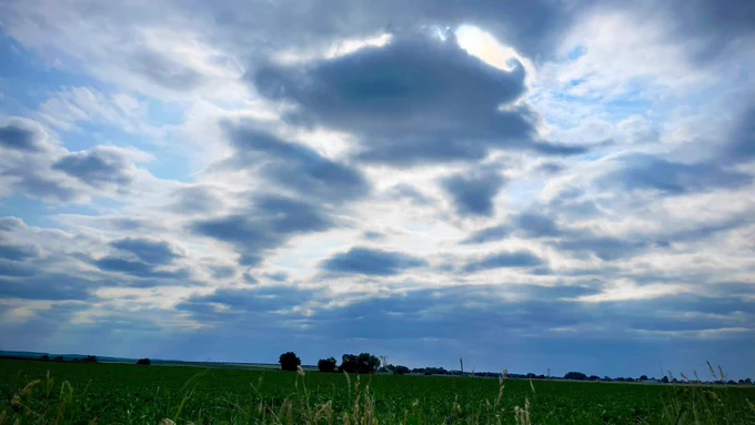 A vast open field under a sky filled with dramatic, textured clouds. Sunlight peeks through, casting a soft glow on the horizon. The scene captures a moody yet peaceful atmosphere.