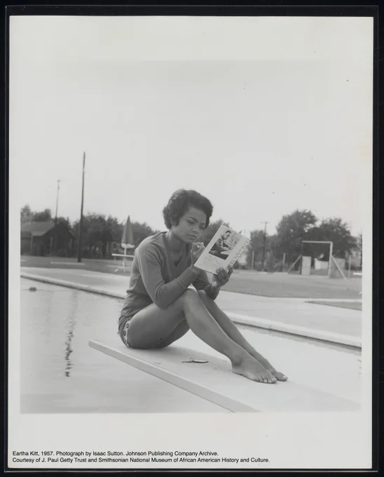 A black-and-white photograph of Eartha Kitt sitting on a diving board above a pool.She holds a copy of the book “Pagan Spain” by Richard Wright. On the board below her is a keychain with a single key. She wears a solid colored long sleeve shirt and short swim bottoms with her legs and feet showing.