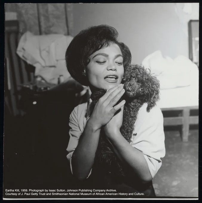 A black-and-white square photograph. Eartha Kitt is center of frame with a fluffy dark colored puppy on her left shoulder. The dog looks directly into the camera with big puppy eyes. Eartha holds up both hands up to cradle the dog on her chest. She wears a light colored elbow length shirt. She is looking down and to the left towards the dog; her eyeliner and lipstick-lined mouth, slightly parted to show teeth, are visible. Her hair is worn short and styled neatly with volume.