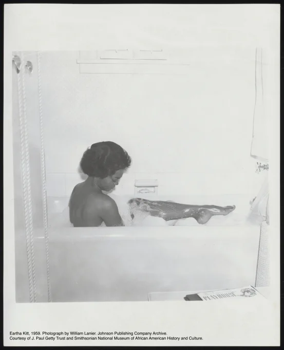 A black-and-white photograph of Eartha Kitt in a bath tub taking a bubble bath. Her back faces the camera while she holds up her leg which is visible from her knee to the tips of her toes. She looks down towards her leg. She is surrounded by white bubbles from the bath. There is a Playhouse magazine in the bottom right foreground of the photo.