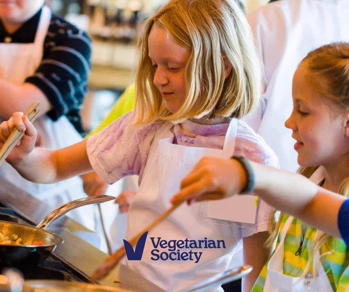 Some children cooking in a class wearing white aprons. The Vegetarian Society is in blue in the bottom middle.