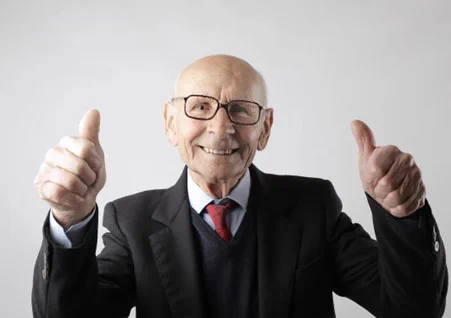 old man wearing glasses, and a dark suit with a red tie, with his thumbs up and smiling