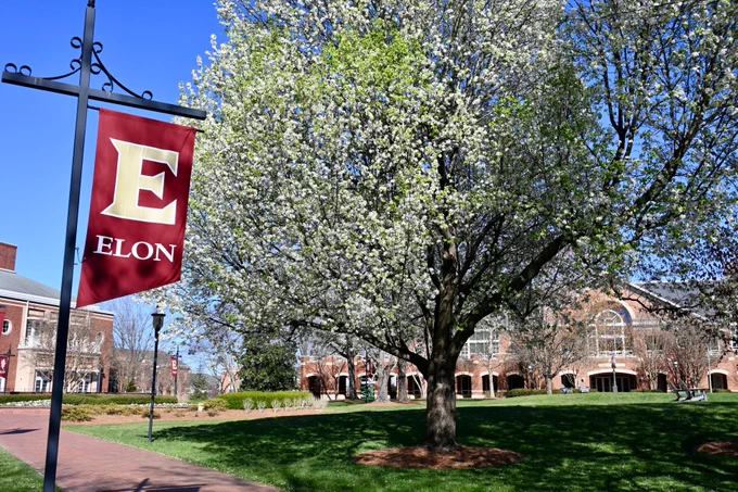 A vibrant scene at Elon University, featuring a burgeonung tree in full bloom beside a campus banner with the letter "E." In the background, traditional brick university buildings are visible under a clear blue sky.