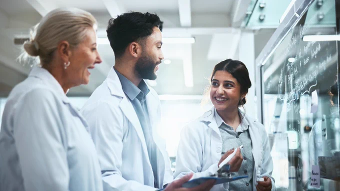 Three people wearing lab coats and talking to each other while writing on a whiteboard