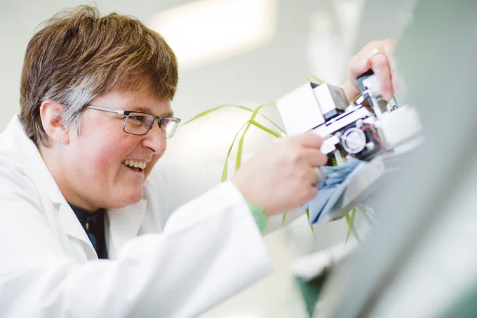 A woman in a lab coat smiles while taking a measurement of leaves with a device.