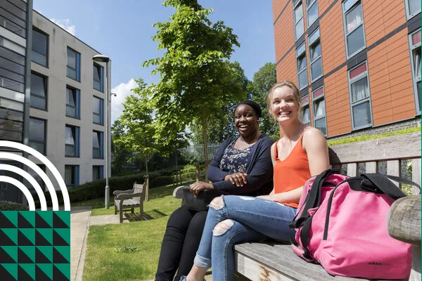 Two female students sat on bench in sunshine outside Horton Halls in Tooting