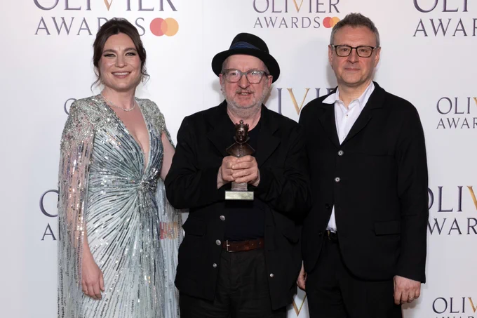 Three individuals posing at the Olivier Awards, with the person in the centre holding an award statue.
