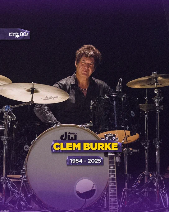An image featuring Clem Burke seated at a drum set with his name and the years 1954 - 2025 displayed on the bass drum. He is surrounded by cymbals and is lit up under dim stage lighting.