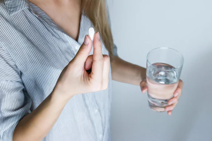 Lady holding glass of water in one hand and a white pill in another. 