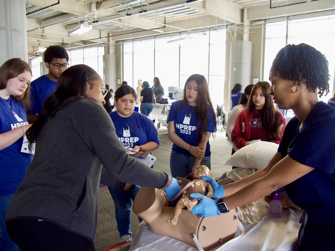 A group of students observe a demonstration on a mannequin led by two instructors in a classroom setting.