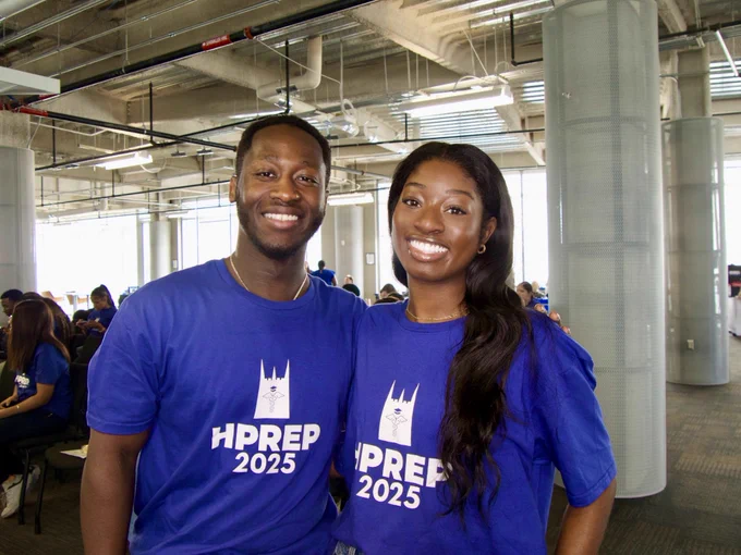 Two individuals wearing blue "HPREP 2025" t-shirts smiling in an indoor setting with others in the background.