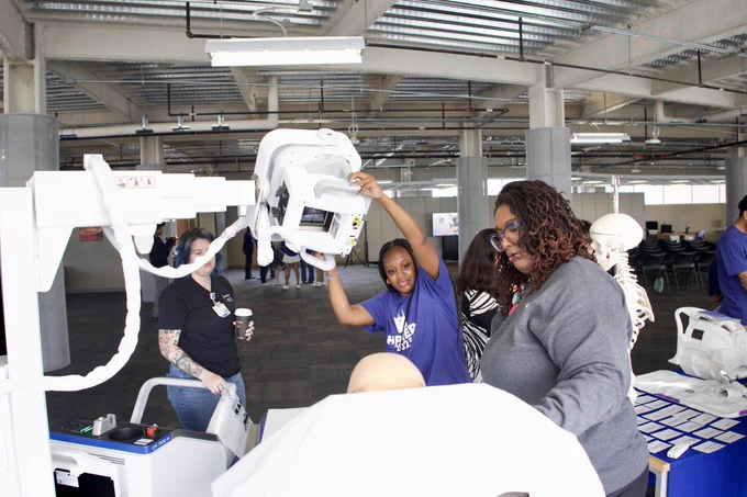 Three individuals interacting with medical training equipment in a spacious room with visible structural elements on the ceiling. 