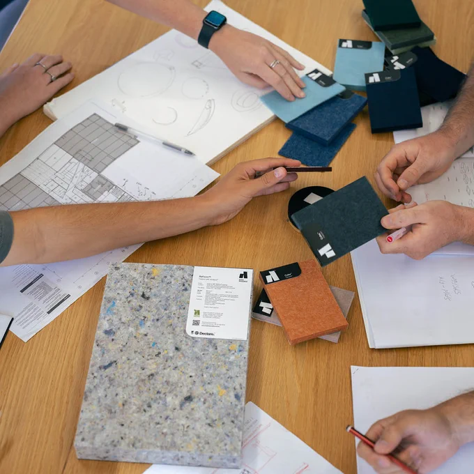 Several people at a table discussing and reviewing material samples and design sketches, including papers and fabric swatches.