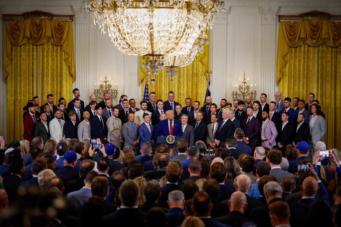A group photo of the Los Angeles Dodgers all dressed in various suits and ties standing inside the White House. President Donald Trump stands at a podium in front of them. The World Series trophy is visible to the left of the podium.