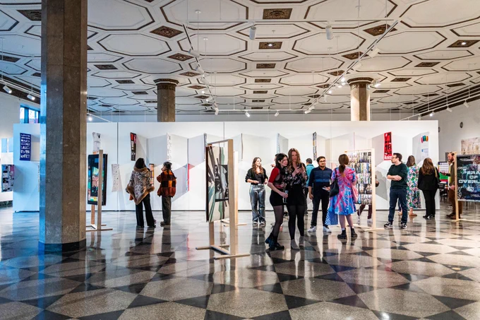 Visitors mingle and view artwork at a Boston University MFA Graphic Design reception in the spacious, well-lit 808 Gallery with a patterned tile floor and ornate ceiling. Freestanding panels display colorful, contemporary graphic pieces, and attendees engage in conversation and observe the work.