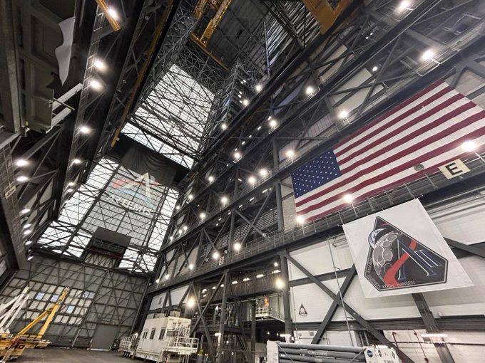 A wide angle image of the new Artemis II mission patch banner hanging in the Vehicle Assembly Building transfer aisle. An american flag hangs above it, while a banner with the Artemis logo is seen hanging at the end of the aisle. 