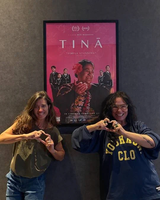 Rachel House and friend stand infront of a Tinā poster making heart fingers
