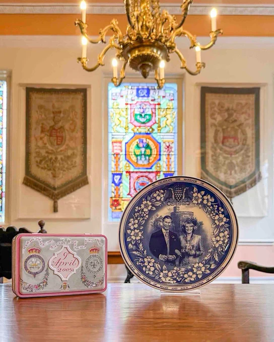 Commemorative tin and plate resting on a table with tapestries, stained glass in the background.