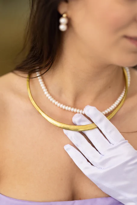 Cropped image of model wearing a shiny gold choker that sites around her collarbone. She has layered it with a white pearl necklace that sits inside the choker. The model is wearing a white glove with her hand placed the choker.