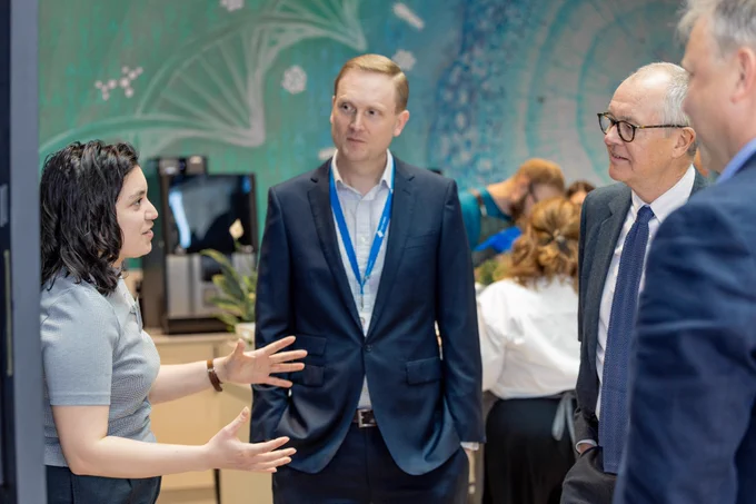 Three men — David Hulcoop, Minister Patrick Vallance, and Ewan Birney — listen with interest to a Helena Cornu, a woman who is talking animatedly. They are seen in profile, dressed formally. Other people can be seen in the background. 