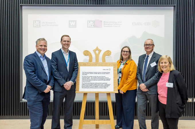 Five people dressed officially stand around a plaque on a stand, smiling at the camera; two men on the left and a man and two women on the right. The slide projected behind them features logos from UKRI, Wellcome, and EMBL-EBI. From left to right: Ewan Birney, David Hulcoop, Jo McEntyre, Patrick Vallance, and Janet Thornton.