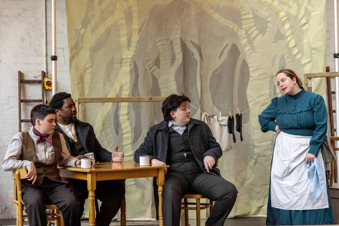 Four actors performing around a table, one standing and three seated.