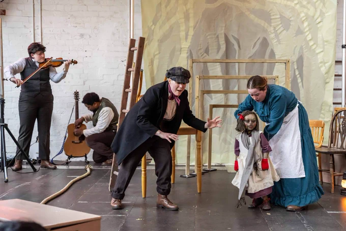 Two actors at the rear of the stage playing instruments. Two actors in the foreground interacting with the puppet of a young girl.