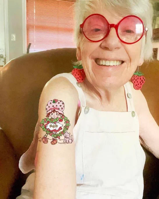 Image of a little old white lady wearing red strawberry earrings, matching red circle framed glasses and a white pair of overalls. There is a tattoo of Strawberry Shortcake on her arm. She’s sitting in front of a brown chair.