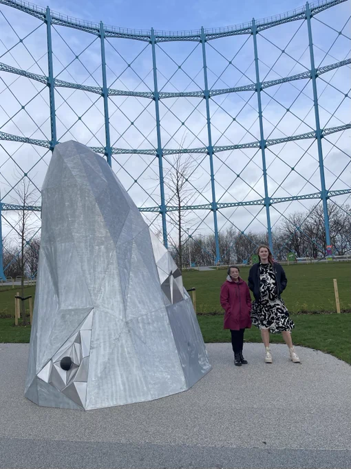 Two individuals standing next to a large, geometric metal sculpture at the base of the Gasholder structure.