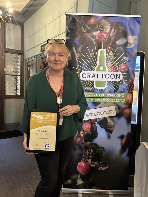 Image of Alison Taffs with her certificate awarding her the outstanding contribution to cider culture, the inaugural Susanna Forbes award. Alison is standing in front of a CraftCon banner which features images of apples and the CraftCon logo, with text in white with a green background underneath. The logo is also a green bottle with the words CraftCon in front in black text.