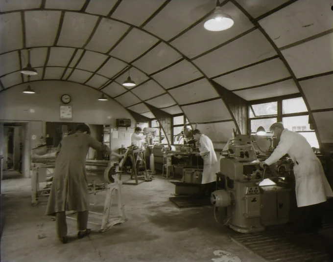 Black and white image of a group of men in lab coats working at various equipment in a room with an oval shaped roof.
