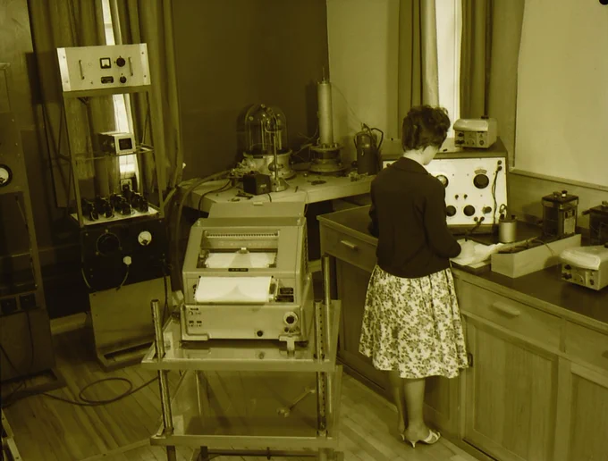 A woman in a cardigan and flowery skirt working at a bench in a room with equipment behind her.