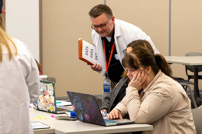 Three educators at CuriousCon. One individual wearing a lab coat and holding a clipboard is interacting with two others seated at a table with laptops and notes.