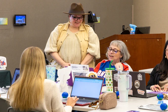Three educators around a table filled with books, mugs, and various office supplies are engaged in conversation. One person is standing and wearing a hat, while the other two are seated, visibly engaged, and smiling during the interaction.