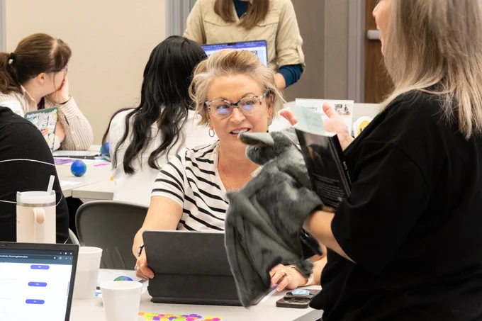 Educators sit at tables, discuss, and work on laptops. One person is holding a bat toy while discussing STEAM career paths with another across the table.