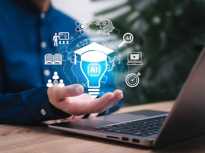 A person's hand open in front a laptop holding an illustration of a lightbulb and other icons related to AI and learning (Photo by Ticha/stock.adobe.com)
