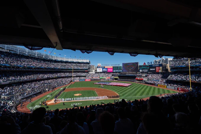 A scenic photo of Yankee Stadium with players lined up on the foul lines and fans standing. A giant American Flag can be seen in the outfield.