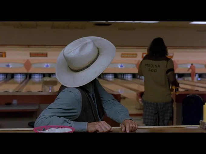 Seated at the bowling alley bar, the Stranger has turned away as he ends his conversation with the Dude, who is walking back to his lane in the background.
