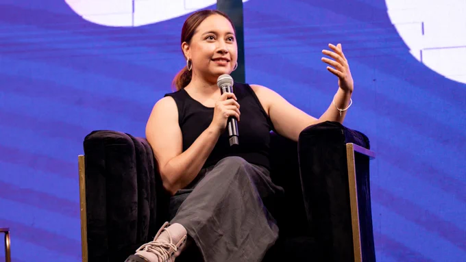 Katya Echazarreta sentada en un sillón negro hablando al micrófono durante el evento "M En la Vida y en la Cancha", con fondo azul y expresión entusiasta.
