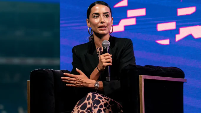 Cristina Llorens, periodista, maestra de yoga y CEO de F45 Sevilla, hablando al micrófono en el escenario del evento "M En la Vida y en la Cancha", con expresión seria y fondo colorido.