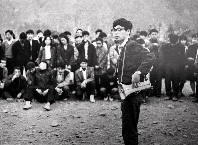 Black and white photo of a young Hayao Miyazaki organizing as chief secretary of Toei's labor union in 1964. he has a bullhorn over his shoulder and there are a number of people in the background.