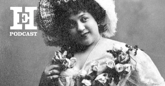 A black and white photo of Belle Elmore, wearing a floral applique dress and large hat. She is looking off camera to the right, and the HistoryExtra logo is in the corner.
