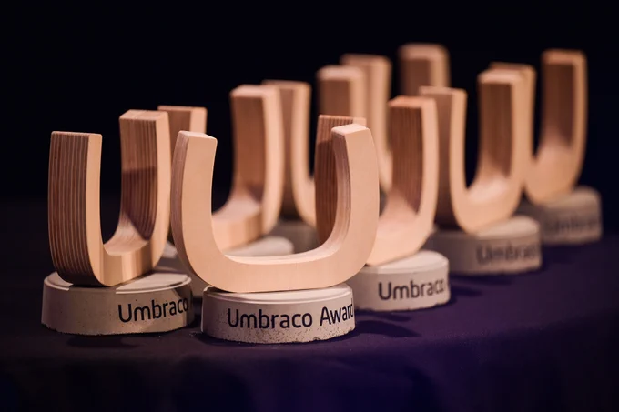 A series of elegant, wooden 'U'-shaped trophies on display, each resting on a gray cylindrical base labeled 'Umbraco Award', against a dark background.