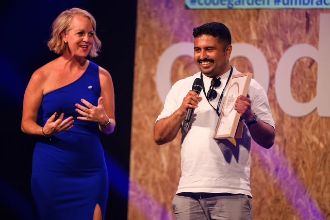 A woman in a blue dress and a man in a white shirt on stage at an event, smiling and interacting. The man is holding a microphone and an award, with a backdrop displaying the event's hashtag and a large 'U' logo.