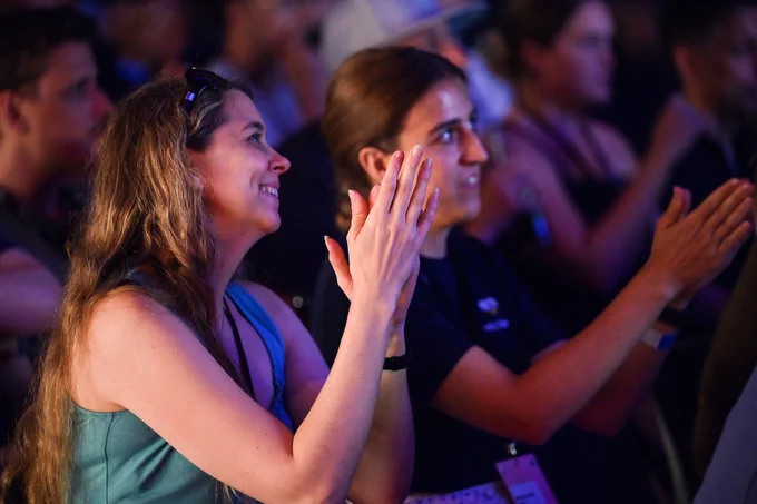 Two women in an audience clapping enthusiastically. The woman on the left has long, wavy hair and is wearing a teal tank top with sunglasses on her head. The woman on the right, in a black t-shirt, is focused on the stage with a joyful expression.