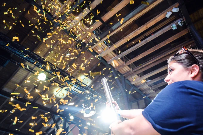 A woman in a blue t-shirt firing a confetti cannon indoors, creating a vibrant shower of golden confetti pieces that cascade through the air under a wooden beam ceiling.