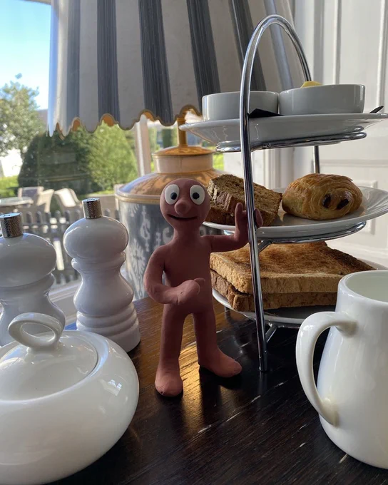Image of Morph  standing on a table with toast, pastries, and tea set, near a window with curtains.